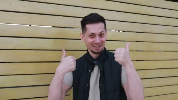 Een vrolijke jongeman in een casual shirt en vest staat voor een houten lattenwand en geeft een dubbele duim met een heldere, zelfverzekerde glimlach. - Video