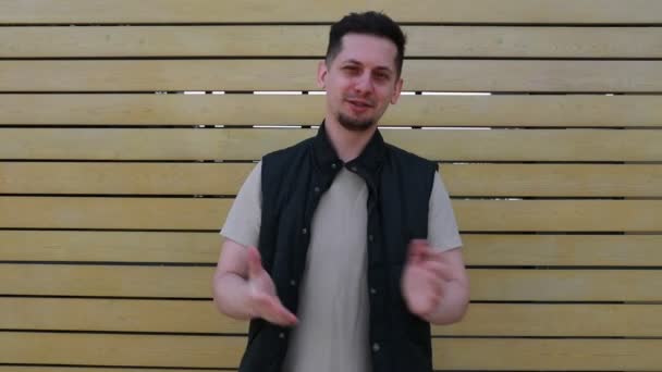 A cheerful young man with short hair, dressed in a sleeveless jacket over a white shirt, claps his hands together while standing in front of a wooden slated wall. His upbeat gesture and casual style - Footage, Video