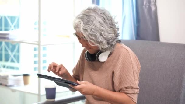 Starsza Azjatka używa tabletu do pisania płatności online, grania w gry, mediów społecznościowych, sprawdzania poczty e-mail, siedzenia na kanapie w salonie. - Materiał filmowy, wideo