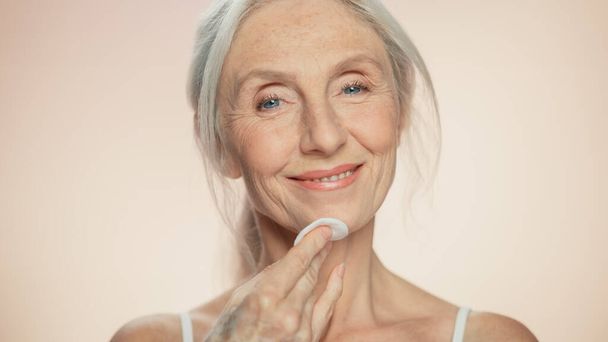 Medium Potrait: Beautiful Senior Woman Uses Cotton Wool Pad to Clean Her Perfect Face of Cosmetics. Smiling Elderly Lady with Soft Skin. Graceful Old - Zdjęcie, obraz