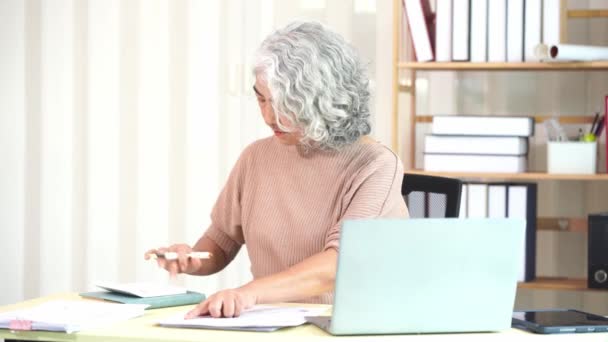 Ältere Asiatin erledigt Papierkram am Computer-Schreibtisch, während sie zu Hause ist Seriöses Gesicht und Stapel von Dokumenten auf dem Tisch, Konzept für die Heimarbeit. - Filmmaterial, Video