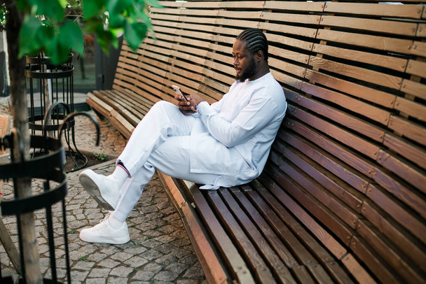 Αφρο-Αμερικανός hipster τύπος σε πολιτιστικά και εθνοτικά ρούχα dashiki κουβέντα σε smartphone σε εξωτερικούς χώρους, γραπτών μηνυμάτων με τους φίλους στα κοινωνικά δίκτυα. Millennial και έννοια του Διαδικτύου. - Φωτογραφία, εικόνα