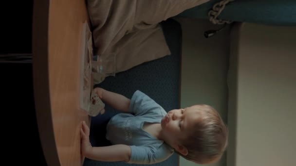 空腹の幼児はキッチンスタジオの丸テーブルの近くの快適なソファーに座ってスナックを食べます. かわいい小さな金髪の少年は朝食の遅い動きを楽しみます 垂直映像. - 映像、動画