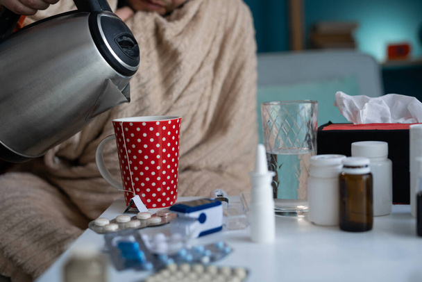 Άρρωστος άντρας με κουβέρτα που φτιάχνει τσάι ενώ κάθεται στον καναπέ. Το άτομο που αισθάνεται άσχημα έχει ιατρική θεραπεία για τα συμπτώματα της γρίπης. Ρίχνει ζεστό νερό σε κούπα - Φωτογραφία, εικόνα