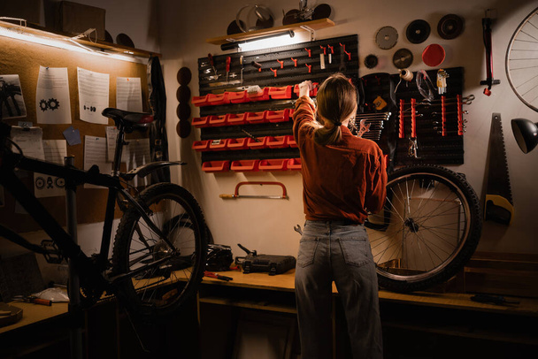 ワークショップの道具で壁の近くに立っている若い女性のリアビュー. 自転車修理とメンテナンスのコンセプト。 コピースペース - 写真・画像
