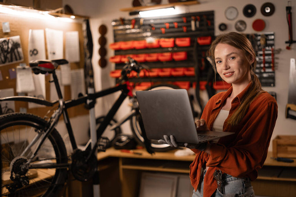 Жінка-механік у своїй майстерні або гаражі дивиться на ноутбук. Власник велосипедного магазину з ноутбуком. Копіювати простір - Фото, зображення