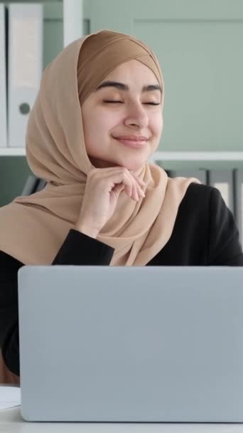Retrato de una alegre mujer de negocios árabe usando hijab, soñando despierto en la oficina moderna. Mujer feliz y satisfecha durante el descanso laboral. Vídeo vertical. - Imágenes, Vídeo