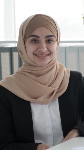 Retrato de una alegre y feliz empresaria árabe con un hiyab en la oficina moderna. Satisfecho mirando a la cámara y sonriendo. Vídeo vertical. - Metraje, vídeo