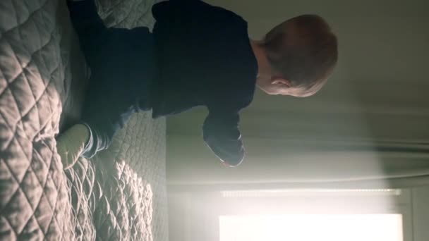 Glückliches blondes Kleinkind springt auf weiches Bett und schaut aus dem hellen Schlafzimmerfenster. Kleiner Junge genießt sonnigen Morgen im Ferienhaus am Wochenende Zeitlupe. - Filmmaterial, Video