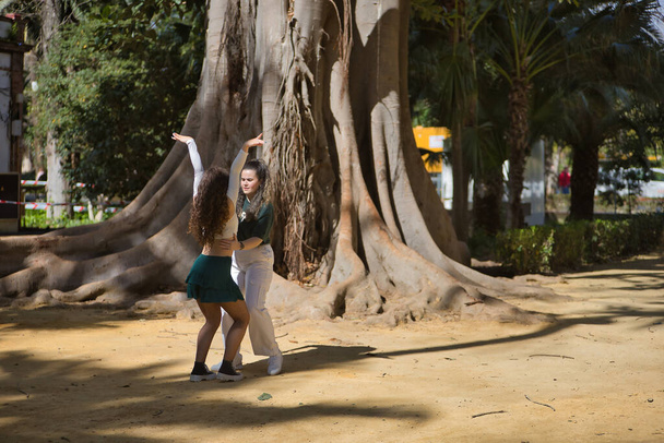 Latein-Frauen-Paar, jung, tanzen Bachata mit einem großen Baum im Hintergrund in einem Outdoor-Park und führen verschiedene Tanzfiguren auf. Konzepttänze, urban, latein. - Foto, Bild