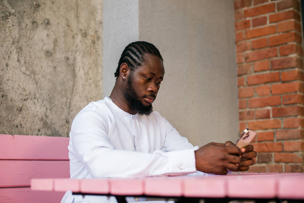 Afrykański Amerykanin w stylowych etnicznych dashiki nosić oglądanie zdjęć w sieciach społecznościowych na telefon komórkowy korzystających z wolnego czasu na świeżym powietrzu. Ciemnoskóry mężczyzna ogląda śmieszne filmy na smartfonie i portalach społecznościowych - Zdjęcie, obraz