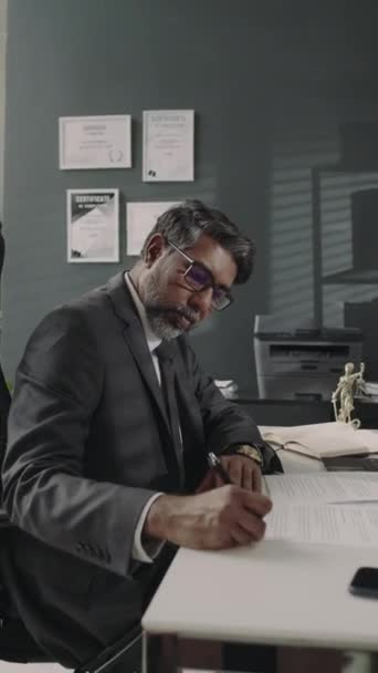 Vertical side footage of busy Biracial adwokat w okularach i garniturze notatki w papierkowej robocie podczas pracy w biurze w agencji pomocy prawnej - Materiał filmowy, wideo