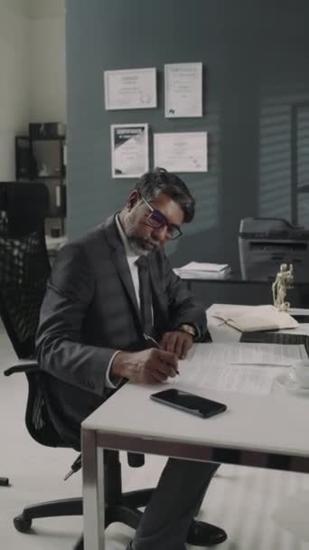 Вертикальные кадры серьезного адвоката-бирасиала в дорогом костюме и очках, работающего с документами за столом в современном офисе в юридической фирме - Кадры, видео
