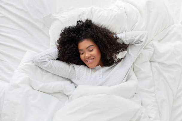 Mujer hispana sonriente está acostada en una cama con sábanas blancas crujientes, sus ojos cerrados en la relajación. La habitación es tranquila y minimalista, con luz natural suave filtrándose a través de una ventana. - Foto, imagen