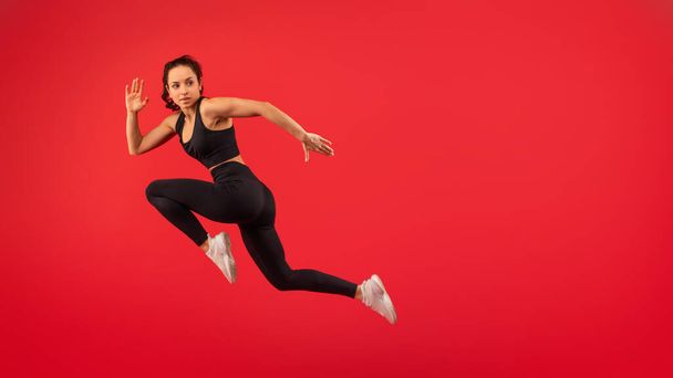 Una mujer que lleva un top negro y polainas negras es capturada en medio del salto en el aire. Sus brazos están extendidos y sus piernas dobladas, mostrando su atletismo y agilidad mientras desafía la gravedad. - Foto, imagen