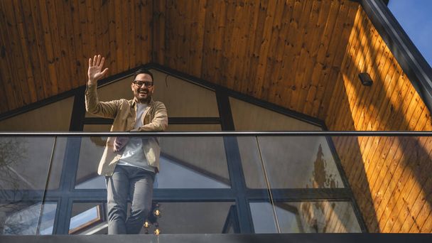 Adult caucasian man with eyeglasses stand on the balcony waving and say hello - Photo, Image