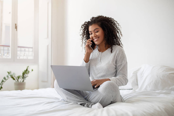 Latynoska kobieta siedzi na łóżku, wielozadaniowa rozmawiając przez komórkę podczas korzystania z laptopa. Wydaje się skupiona i zaangażowana w swoje działania, równoważąc oba urządzenia wygodnie. - Zdjęcie, obraz