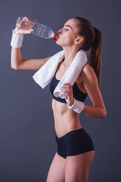 Beautiful girl in sportswear is holding a towel and drinking water, on gray background - Photo, Image