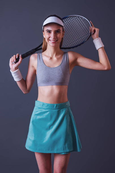Beautiful girl in sportswear is holding a tennis racket, looking at camera and smiling, on gray background - Zdjęcie, obraz