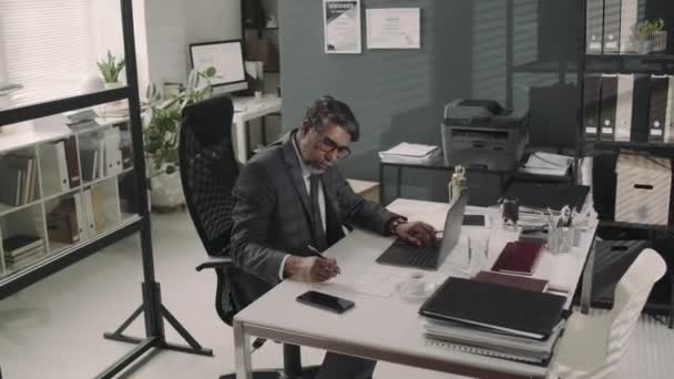 Kaaren kuvamateriaalia mies Biracial asianajaja työskentelee kannettavan tietokoneen ja tehdä muistiinpanoja asiakirjoissa työpäivän aikana moderni asianajotoimisto - Materiaali, video