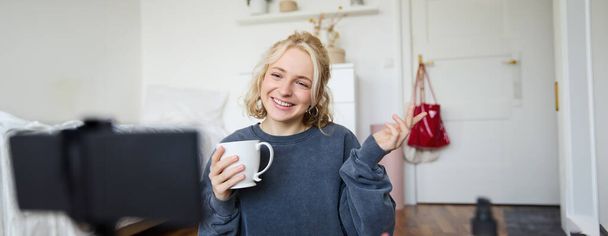 Porträt der süßen lächelnden Frau, blondes Mädchen trinkt Tee und nimmt einen lässigen Lifestyle-Videoblog auf, Vlogger sitzt in einem Raum mit Kamera und Stabilisator, hält eine Tasse Kaffee und spricht. - Foto, Bild