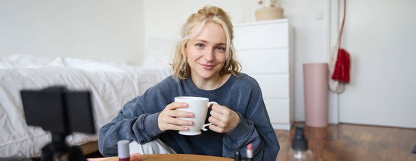 Close-up portret van mooie blonde vrouw, meisje zit in haar kamer, doet make-up, neemt video op voor sociale media, drinkt thee, houdt mok en glimlacht op camera. - Foto, afbeelding