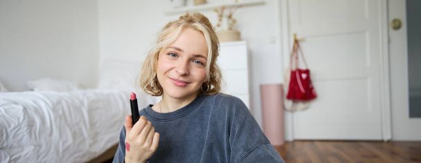 Portret van de jonge make-up kunstenaar, beauty blogger met nieuwe lippenstift, video opnemen in haar kamer, glimlachen en positiviteit uitdrukken. - Foto, afbeelding