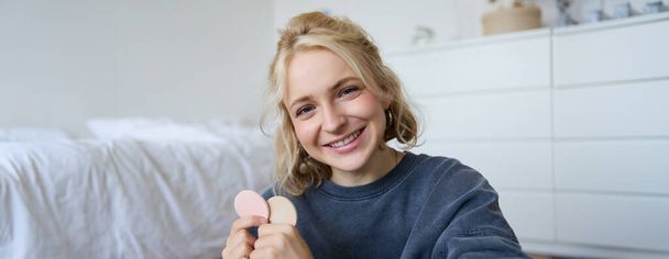 Close up portrait of happy, beautiful young vlogger, content maker recording video about makeup, showing beauty products on camera, smiling happily. - Photo, Image