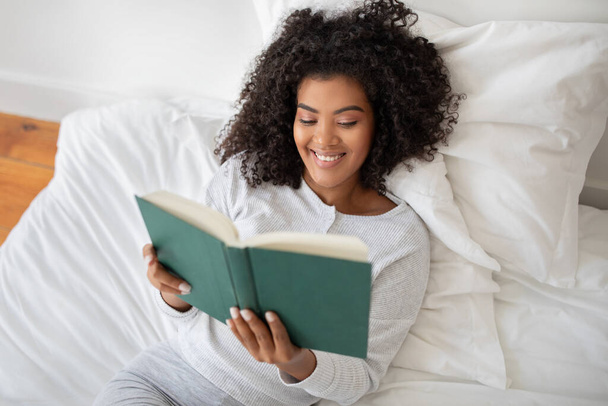 Die hispanische Frau liegt auf einem Bett, in ein Buch vertieft, das sie in ihren Händen hält und lächelt. Der Raum ist schwach beleuchtet, und sie wirkt entspannt und konzentriert auf ihre Lektüre.. - Foto, Bild