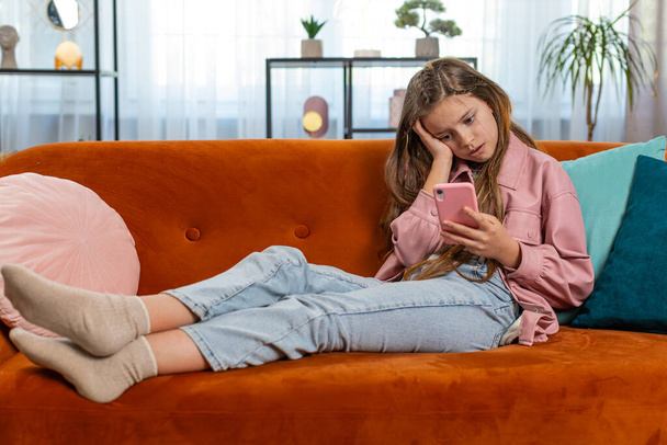 Upset girl using smartphone playing game loses becoming surprised sudden lottery results, bad news, fortune loss, fail education study test. Female teen child kid at home in living room sits on sofa. - Valokuva, kuva
