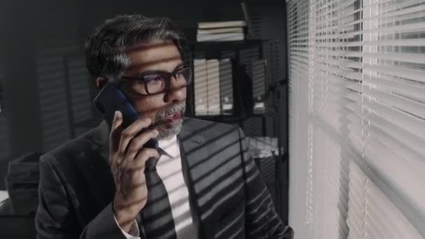 Chest up footage of senior bearded Biracial attorney in suit and glasses speaking on mobile with customers and clients discussing legal issues and peeking through blinds in office - Footage, Video