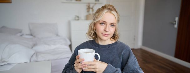 微笑む若い金髪の女性の肖像画を閉じる,寝室で紅茶のカップと座って,自宅で休憩し,彼女のコーヒーを楽しむ. - 写真・画像