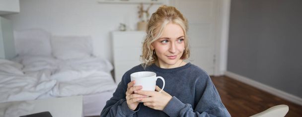 Portret uśmiechniętej pięknej młodej kobiety, siedzącej w sypialni z filiżanką herbaty, odpoczywającej samotnie w domu, cieszy się weekendem w domu. - Zdjęcie, obraz