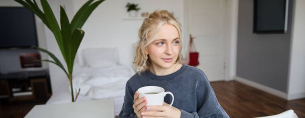 Lähikuva muotokuva hymyilevä nuori vaalea nainen, istuu kupin teetä makuuhuoneessa, lepää kotona, nauttii hänen kahvia. - Valokuva, kuva