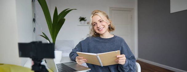 Porträt einer lächelnden blonden Frau, die im Schlafzimmer sitzt, Laptop und Digitalkamera benutzt, Videos für Lifestyleblog aufnimmt, liest, ihr Notizbuch benutzt. - Foto, Bild