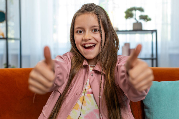 Наприклад. Щаслива молода кавказька дівчинка дивиться схвально на камеру, показуючи подвійні великі пальці вгору, як знак позитивного чогось хорошого, позитивного відгуку. Жінка-підліток вдома у вітальні на дивані. - Фото, зображення