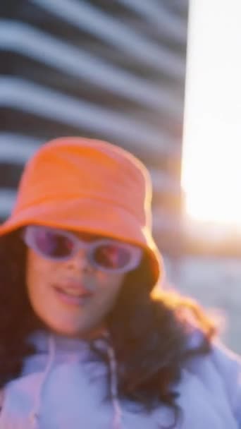 Pionowe wideo. powolny ruch fantazyjne młody brunetka wieloetniczne kobieta noszenie żywe jasnopomarańczowy kapelusz, taniec głupca wokół, zabawy gestykulacji z rąk, korzystających zachód słońca na zewnątrz na miasto - Materiał filmowy, wideo