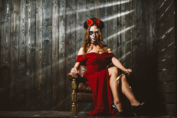 Una mujer cautivadora vestida de rojo con el maquillaje tradicional del Día de los Muertos se sienta con confianza, su pose emana un encanto misterioso. Misteriosa Dama con el Día de los Muertos Maquillaje - Foto, imagen