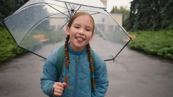 Veselý holčička při pohledu na kameru show jazyk grimasa tvář výraz školačka dítě dcera hýčkat zábava školačka žák dítě šťastný déšť počasí dětinské víkend energetický radovat park město - Záběry, video