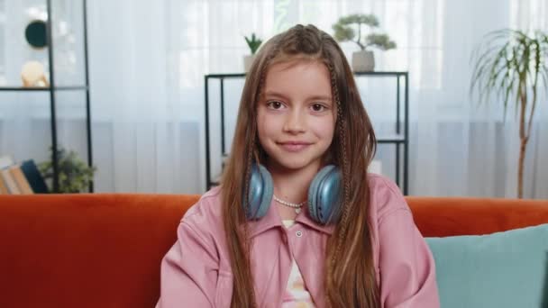Portrét šťastné mladé dívky s sluchátky kolem krku sedí na pohodlné pohovce a dívá se do kamery v obývacím pokoji v bytě. Roztomilé puberťačky s dlouhými hnědými vlasy. Životní styl. - Záběry, video