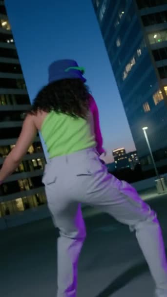 Vertical video. Sexy cool girl staring at camera at night downtown background. Beautiful mixed race woman clubbing at disco party on rooftop. Portrait of charming female person dancing on neon city - Footage, Video