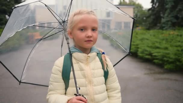 Маленька європейська дівчинка дивиться на камеру, посміхається, тримає парасольку, дощова погода, дитина, зіниця, місто поза парком, виражає смішний дитячий парасольковий рюкзак, дитячий садок, насолоджуйтесь - Кадри, відео