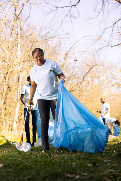 Αφροαμερικάνος εθελοντής τακτοποιεί την περιοχή του δάσους με σκουπίδια και πλαστικά μπουκάλια, μαζεύοντας σκουπίδια με νύχια και τσάντες. Νεαρή γυναίκα που εργάζεται εθελοντικά για τη διατήρηση του φυσικού περιβάλλοντος. - Φωτογραφία, εικόνα