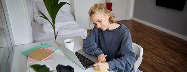Retrato de uma jovem sorridente, estudante do sexo feminino, fazendo curso a distância, usando laptop, estudando em casa, escrevendo, fazendo anotações. - Foto, Imagem