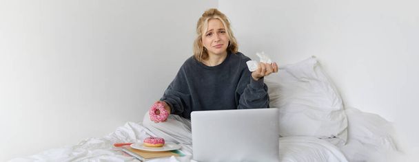 Портрет розчарованої, пригніченої молодої жінки, що плаче, сидить на ліжку з ноутбуком, їсть їжу комфорту, тримає пончик і витирає сльози. - Фото, зображення