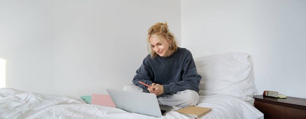 Retrato de una joven sonriente estudiando en su cama, trabajando desde casa en el dormitorio, sentada con portátil y cuadernos en pose de loto, luciendo feliz y relajada. - Foto, Imagen