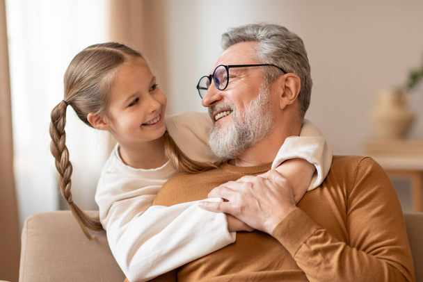 Senior man and a little girl are seated on a couch, looking towards each other, grandfather and granddaughter embracing at home - Photo, Image