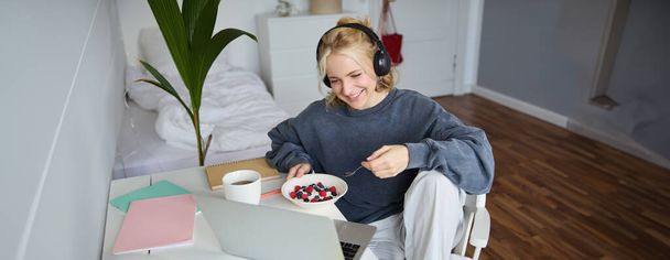 Mosolygó szőke nő portréja fülhallgatóban, ül a szobában, filmet néz laptopon, reggelizik és teát iszik, ebédel a számítógép képernyője előtt. - Fotó, kép