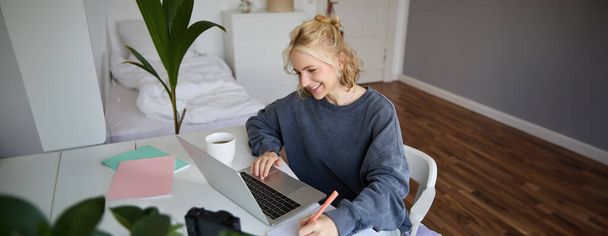 Портрет усміхненої молодої жінки, студентки, яка займається дистанційним навчанням, використовує ноутбук, навчається вдома, записує, робить нотатки. - Фото, зображення