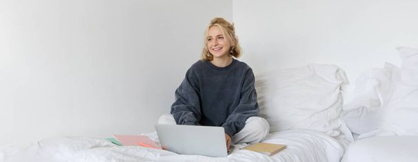Bild einer glücklichen jungen Frau, E-Learning-Studentin von zu Hause aus, Verbindung zum Online-Kurs auf ihrem Laptop, sitzt mit Notizbüchern im Bett, lächelt und sieht glücklich aus. - Foto, Bild